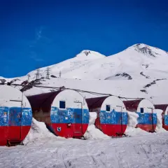 Białe, beczkowate schroniska z malowanymi flagami Rosji, zasypane śniegiem, na tle ośnieżonego Elbrusa. Wejście na szczyt.