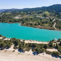 Turkusowa laguna na Kassandrze, otoczona zielonymi wzgórzami i sosnowym lasem. W oddali widać zabudowania i plażę z łodziami.