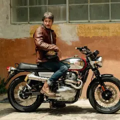 Uomo con giacca in pelle marrone seduto su una moto Royal Enfield Interceptor. Il manubrio cafe racer è cromato, pronto per un viaggio.