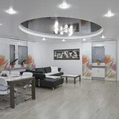 Nowoczesny salon z okrągłym sufitem napinanym, który odbija elegancki żyrandol. Wygodna kanapa i stół z krzesłami tworzą przytulną przestrzeń.