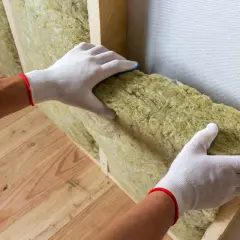Mains gantées installant un isolant minéral dans une structure en bois pour une isolation intérieure efficace.