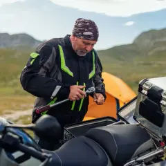 Hombre con chaqueta de moto repara su vehículo, quizás para evitar que la moto se ahogue. Montañas y tienda de campaña al fondo.
