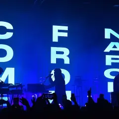 Sul palco, silhouette di musicisti con tastiere e microfoni, mentre sullo sfondo campeggia la scritta "COM FROM NAPOLI" in lettere luminose.