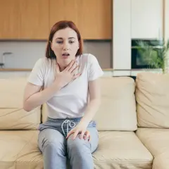 Une jeune femme assise sur un canapé, la main sur la poitrine, le souffle court. Elle semble avoir du mal à parler.