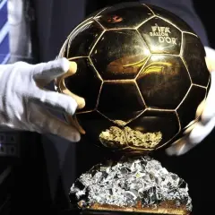 Złota Piłka FIFA Ballon d'Or na błyszczącym cokole, trzymana w białych rękawiczkach.