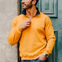 Un homme ethnic blue porte un pull jaune vif avec un col bleu marine, ajustant la fermeture éclair. Il est appuyé contre un mur en pierre.