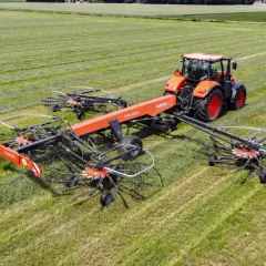 Pomarańczowy traktor zgrabnie manewruje po polu, ciągnąc szeroką zgrabiarkę do traktorka, która sprawnie formuje pokosy zielonki.