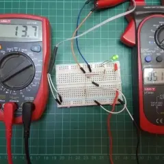Układ na płytce stykowej z diodą Zenera, dwoma multimetrami i diodami LED. Jeden multimetr pokazuje 13.7V, drugi 05.16.