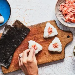Making onigiri rice recipe: hands shaping rice balls with salmon filling, nori sheets, and bowls of rice and salmon.