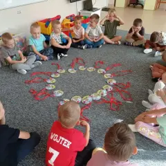 Dzieci siedzą w kółku na dywanie, patrząc na okrąg z zawieszkami. Może to być część zabawy, jak w "Mam chusteczkę haftowaną".