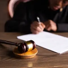 Młotek sędziowski na drewnianym stole, w tle osoba pisząca.