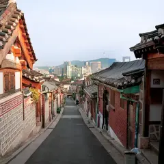 En smal gata kantad av traditionella hus med tegeltak leder mot en modern stadssilhuett i Seoul, Sydkorea.