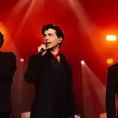 Il trio delle meraviglie, tre cantanti in abiti eleganti, si esibiscono sul palco illuminato da luci rosse.