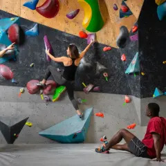 Drei Personen bouldern an einer bunten Wand. Ein Mann gibt Tipps, eine Frau klettert, ein Mann ruht.