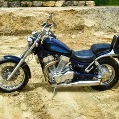 Błękitny motocykl Suzuki Intruder z chromowanymi elementami i czarnym siedzeniem stoi na piaszczystym podłożu.