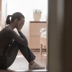 Kobieta siedzi na podłodze, zakrywając twarz dłońmi. W tle widać łóżko i roślinę. To obraz smutku, być może baby blues.