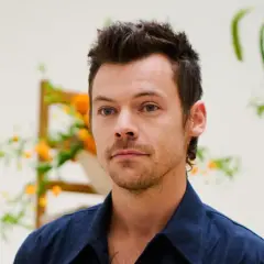 Harry Styles con un moderno mini mullet, mirada intensa y camisa azul, rodeado de arreglos florales.