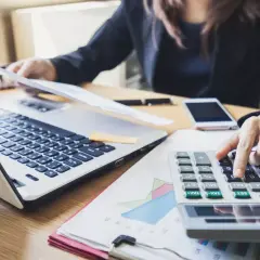 Kobieta w garniturze liczy na kalkulatorze przy laptopie i dokumentach.