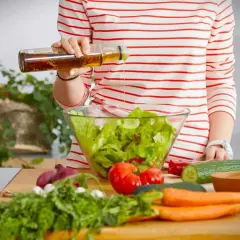 Eine Person bereitet einen Salat zu und träufelt Öl hinein. Frisches Gemüse wie Tomaten, Gurken und Karotten liegen auf dem Tisch. Ein Hauch von weisswein essig rundet das Gericht ab.