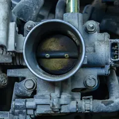 Primer plano de una mariposa de admisión de motor de coche, con suciedad y grasa. Unas manos enguantadas se ven a la izquierda.