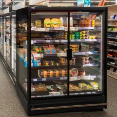Chłodnia z produktami spożywczymi w supermarkecie, z widocznymi serami, wędlinami i deserami.