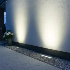 Eleganckie oprawy najazdowe subtelnie oświetlają ścieżkę i białą elewację domu, tworząc nastrojową atmosferę wieczorem.