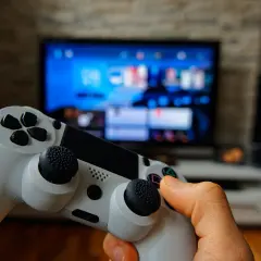 Dłoń trzyma biały kontroler, gotowa do gry na konsoli PS5 Pro. W tle widać rozmazany ekran telewizora z menu.