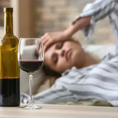 Kobieta leży na kanapie z ręką na czole, wyraźne objawy kaca. Obok butelka i kieliszek czerwonego wina.