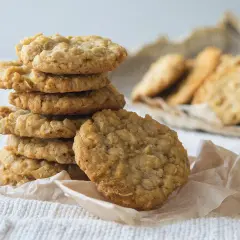 Ein Stapel goldbrauner Haferflocken-Bananen-Cookies auf einem weißen Tuch, daneben weitere Kekse.