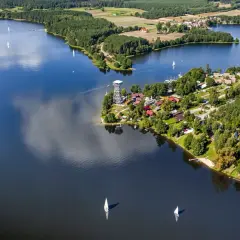 Widok z lotu ptaka na jezioro i zabudowania w Wdzydzach Kaszubskich. Na wodzie żaglówki, a na brzegu wieża widokowa i domki.