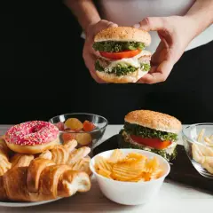 Ręce trzymają burgera, obok talerz z croissantami, pączkiem i ciastkami, miseczki z żelkami, frytkami i chipsami. Czy cheat day przyspiesza metabolizm?