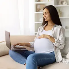 Kobieta w zaawansowanej ciąży sprawdza w internecie informacje o terminie porodu, siedząc na kanapie z laptopem na kolanach.