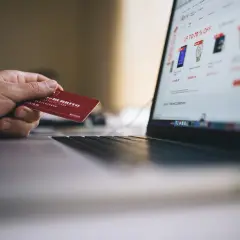 Dłoń trzyma czerwoną kartę przy laptopie z otwartą stroną zakupową. Czy debet jest oprocentowany? To pytanie zadaje sobie wiele osób podczas zakupów online.