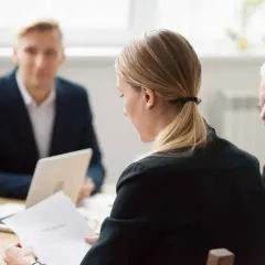 Kobieta z dokumentami w ręku, obok niej siedzi starszy mężczyzna. W tle młody mężczyzna z laptopem. Dyskutują o tym, kim jest prokura.