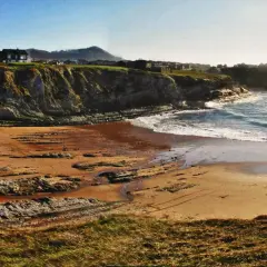 La playa de Portio, con sus acantilados rocosos y arena dorada, se baña en la luz del sol. Las olas rompen suavemente en la costa.