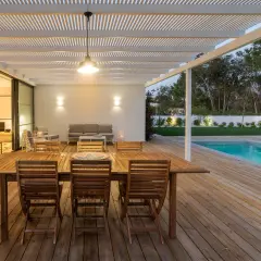 Table et chaises en bois huilé sur une terrasse en bois, avec piscine et jardin en arrière-plan.