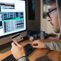 Mężczyzna w kapturze trzyma kartę kredytową, patrząc na ekran z kodem. Zastanawia się, czy bank odda skradzione pieniądze z konta.
