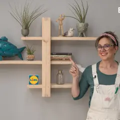 Estantería DIY de madera con pez decorativo, plantas y libros. Una mujer sonríe y señala la estantería.