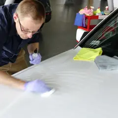 Mężczyzna w rękawiczkach nakłada powłokę ceramiczną na maskę białego samochodu, używając aplikatora i buteleczki z płynem.