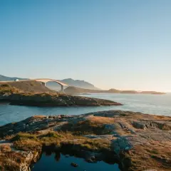 Malownicza norweska zatoka z łukowatym mostem i górami w tle. Słońce zachodzi nad spokojną wodą.