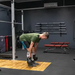 Mężczyzna wykonuje martwy ciąg rumuński z kettlebellami, ćwicząc mięśnie tylnej taśmy. W tle widać sprzęt do treningu siłowego, w tym sztangi i obciążenia.