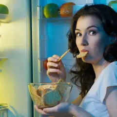 Une femme, prise par une **sensation de faim**, grignote des biscuits devant un réfrigérateur ouvert, rempli de fruits et légumes.