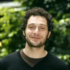 Claudio Santamaria giovane, con capelli ricci e barba, sorride con uno sfondo verde sfocato.