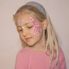 Ein Mädchen mit blonden Haaren und einem Stirnband, das ein Blumenmuster auf der Wange trägt. Das Stern schminken Kinder ist hier als zarte Blüten umgesetzt.