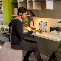 Uomo con cuffie lavora al laptop in un angolo studio in soggiorno. Libri, lampada e pianta completano l'arredo.