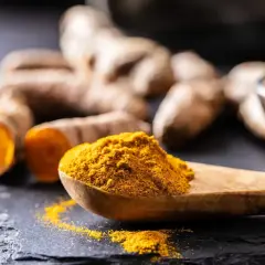 Ein Holzlöffel mit leuchtend gelbem Kurkumapulver liegt neben frischen Kurkumawurzeln.