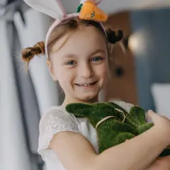 Uśmiechnięta dziewczynka w króliczych uszkach przytula zielonego zajączka. Wesoła wielkanoc w hotelu.