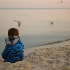 Chłopiec w niebieskiej bluzie siedzi na plaży, obserwując mewy. Spokojna atmosfera nad morzem może pomóc w tym, jak wyciszyć dziecko nadpobudliwe.