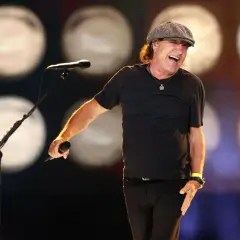 Brian Johnson w czapce i czarnej koszulce śpiewa do mikrofonu na scenie, rozbawiony.