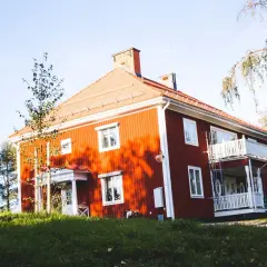 Rött hus med vita knutar och veranda, omgivet av grönska. Ett charmigt vandrarhem i Jokkmokk som bjuder in till en naturnära vistelse.